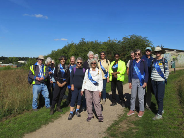 A large group of people, all wearing blue sashes that say u3a, stand on a path through a field. The sky is blue and they are all smiling.