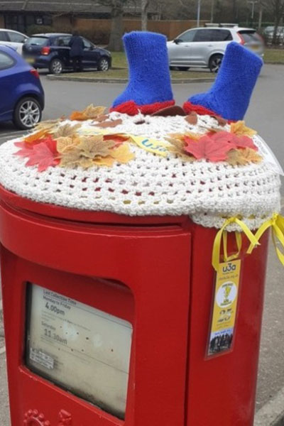 A postbox with a white crochet cover with two blue walking boots on top, and fabric leaves