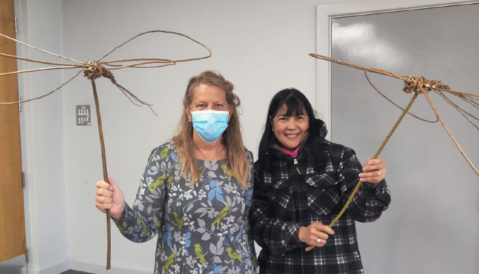 Two women standing next to each other, holding sticks with butterfly shapes at the end - an example of willow weaving.