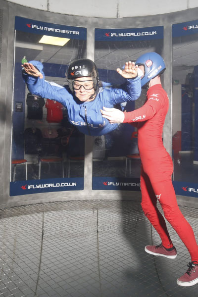 A woman doing indoor sky diving - she is inside a room, her arms are outstretched, it looks like she is flying. A man to the right of her is steadying her.