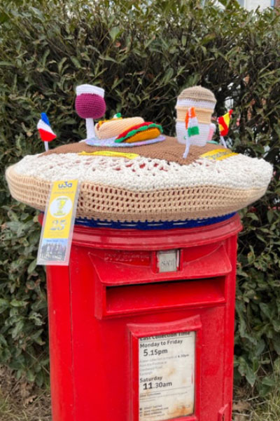 A postbox with a crochet cover with fabric models of a wine glass, various foods and flags