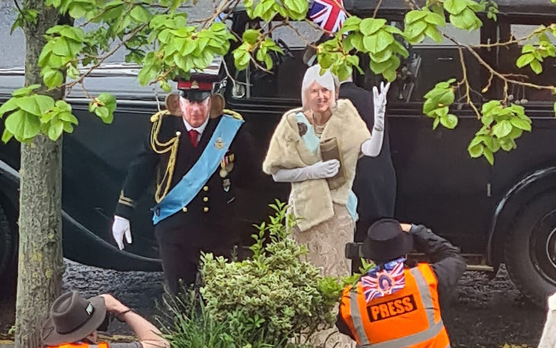 A man and woman wearing face masks depicting King Charles and Queen Camilla step out of a black car and wave.
