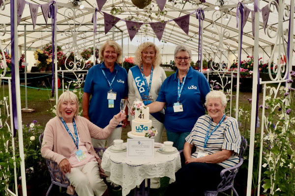 Five women, wearing u3a tshirts sashes and lanyards, inside a marquee. They are inside a large white structure and standing around a table which has a white table cloth and a white cake that says u3a on it. 