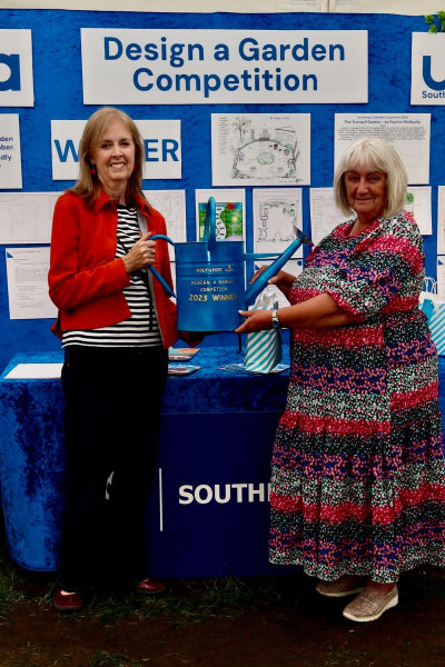 One woman is handing another a watering can that says 'Garden Competition 2023 winner'. Behind them is a display board showing garden designs.