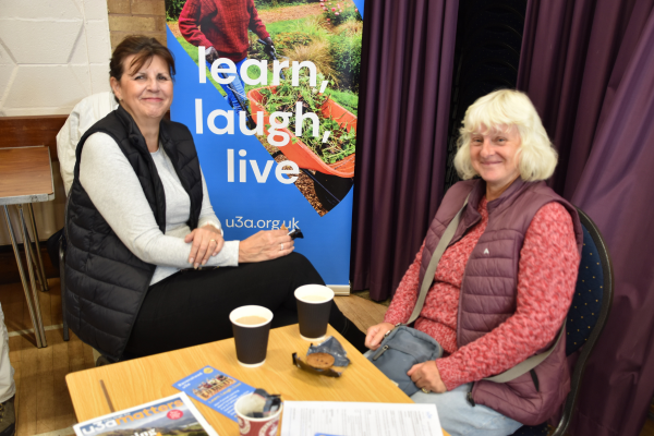 Two women chatting together at a table next to a u3a flag banner that says learn, laugh, live