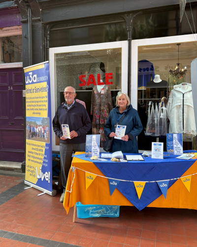 Two u3a members standing by a display of u3a, including a table decorated with u3a bunting and a large u3a banner