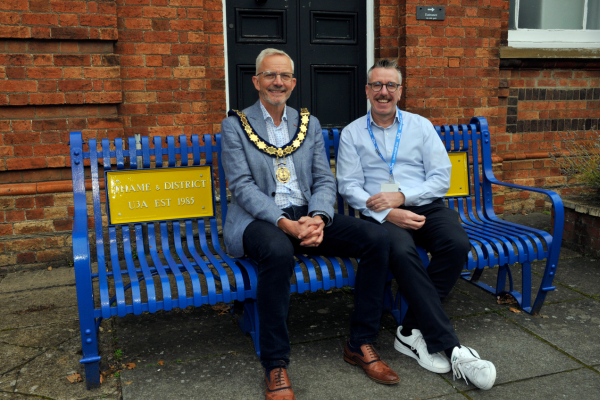 two people sit on a blue and yellow commemorative bench