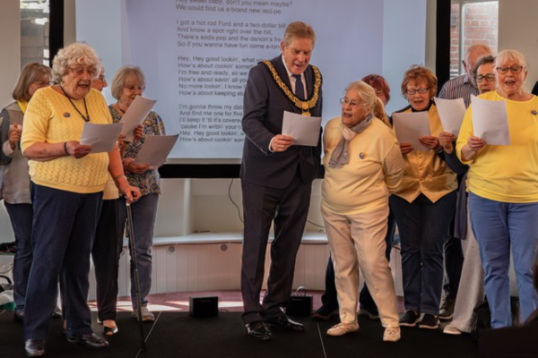 Man in mayor outfit sings from lyric sheet with 10 members of singing group on a stage dressed in yellow and blue clothes